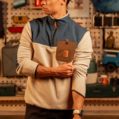 Men's fleece pullover with cream body, navy chest panel, brown pocket detail, and quarter-zip in garage setting.